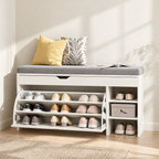 A white entryway shoe bench with a gray cushion, styled with two decorative pillows, an open magazine, and multiple pairs of shoes on open shelves and side storage compartments, set in a bright room with sunlight and a rug.