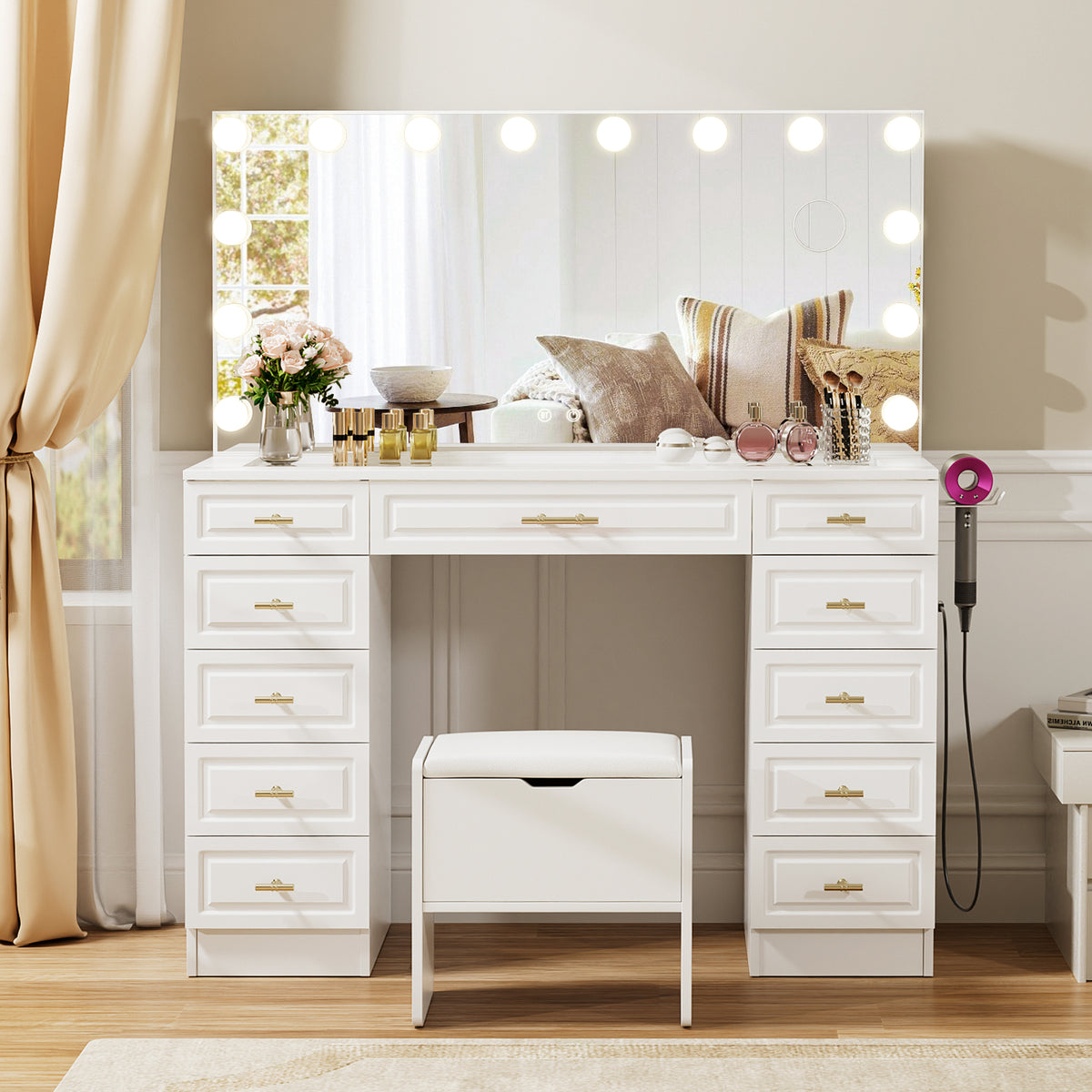 White vanity table with a large lighted mirror, multiple drawers, a matching stool, and gold-tone handles, styled in a bright bedroom or dressing area.